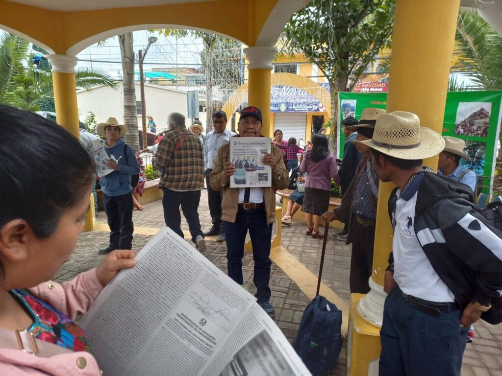 Around 10 community members under a yellow roof. In the foreground, a female-presenting person is reading a copy of the Official Journal of Central America. A male-presenting person is holding up a copy of the Official Journal of Central America
