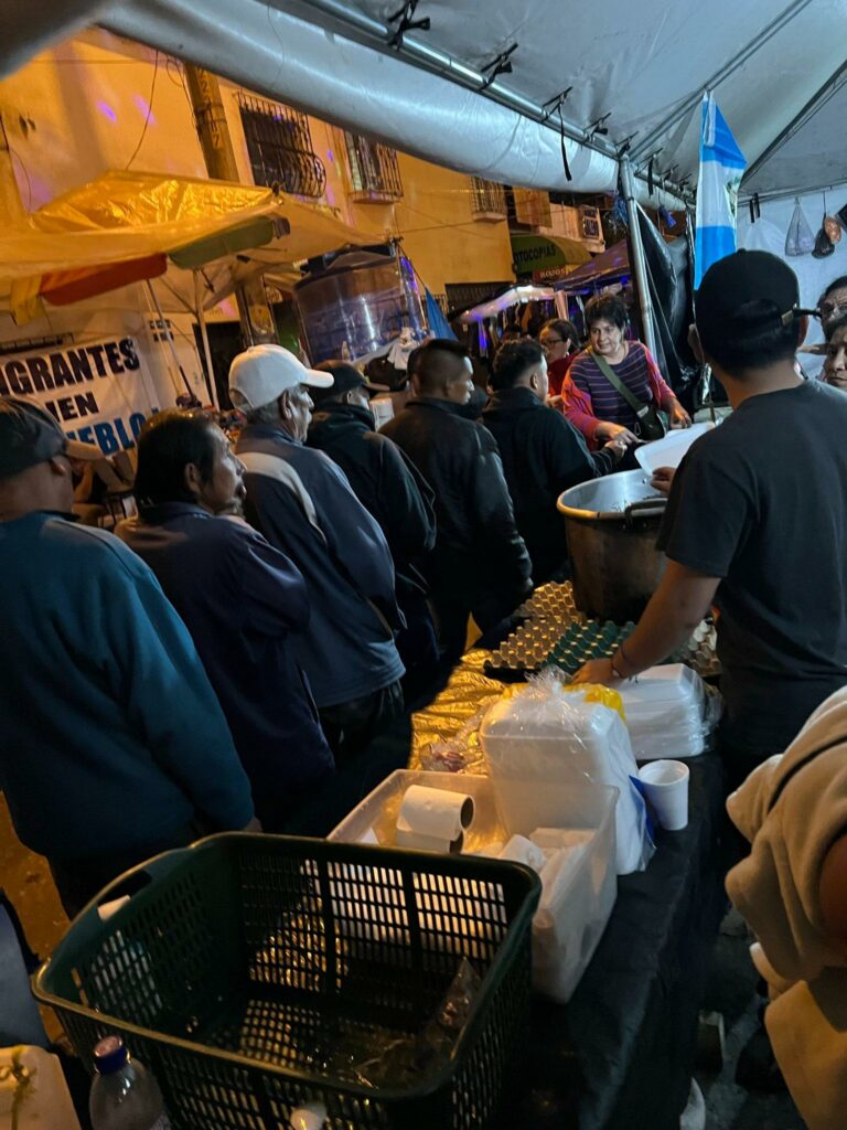 Under a white tent, a line of people wait for food to be served.