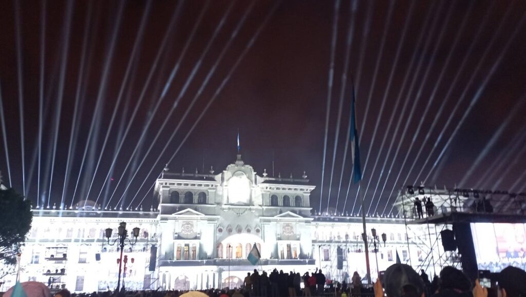 La imagen es una fotografía tomada de noche del palacio nacional de Guatemala, que se encuentra iluminado con luces artificiales y se alcanzan a ver las sombras de personas que están mirando hacia el edificio.