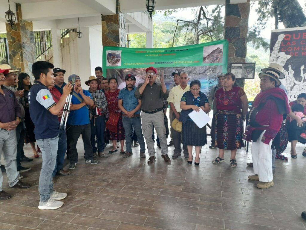 22 personas de pie en semicírculo escuchando al líder de ADH que habla en un mic. Las mujeres se visten con ropa Maya tradicionál. Unos de los hombres llevan sombreros de paja. Al fondo hay un póster verde. Un reportero está de pie hacia el frente.