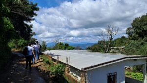A la izquierda, tres personas caminan en una fila en un camino de tierra sombreado por árboles. A su derecha hay una casa blanca con un techo de lámina y en el fondo hay un cielo azul con nubes blancas.